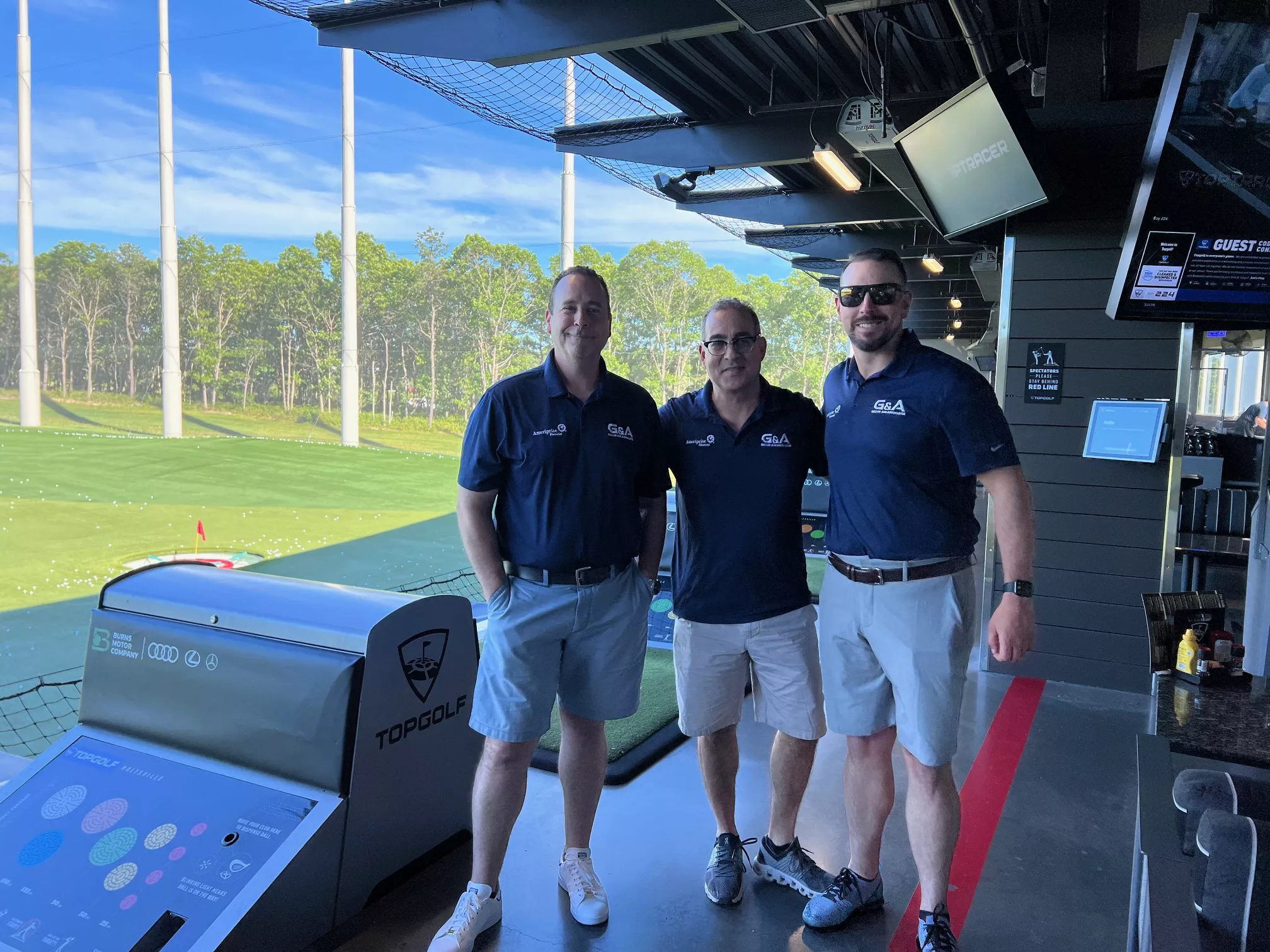 Three men in matching navy polo shirts at a Topgolf driving range bay