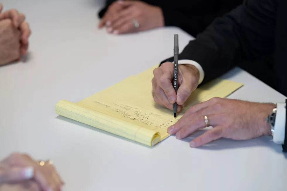 Close-up of a hand writing on a yellow legal pad at a meeting table