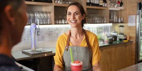 Smiling café owner in an apron and yellow top serving a customer at the counter