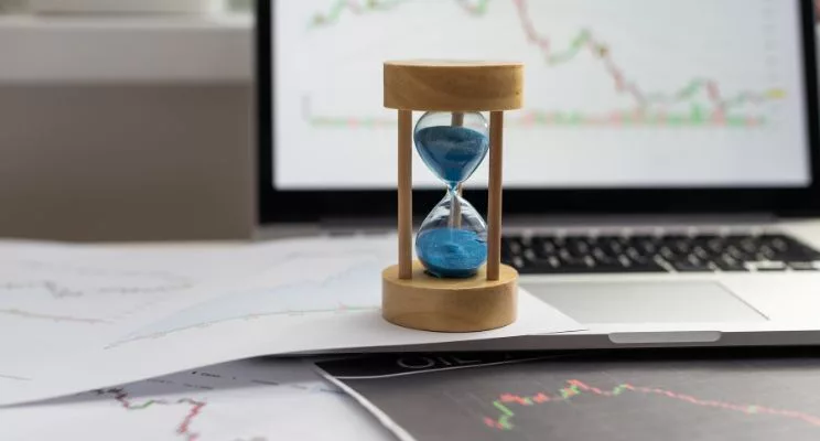Hourglass with blue sand on printed market charts in front of a laptop showing a stock chart