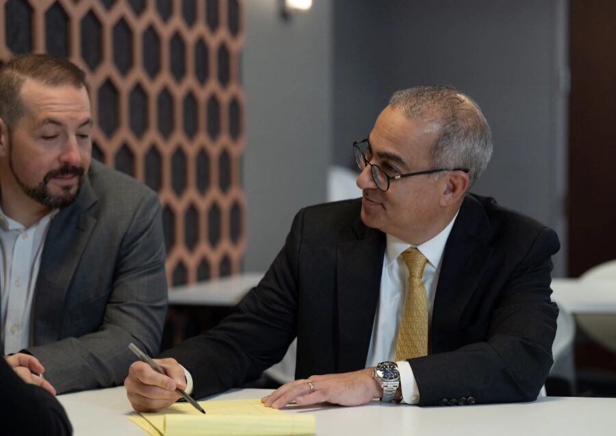 Two advisors in suits at a meeting table, one taking notes on a legal pad